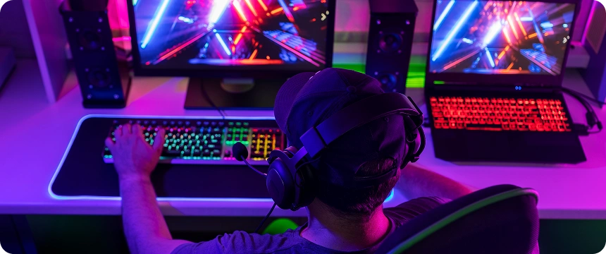 Gracz pogrążony w rozgrywce przy biurku z żywym oświetleniem RGB. Dwa ekrany wyświetlają kolorową scenę akcji. Nastrój emanuje skupieniem i ekscytacją.